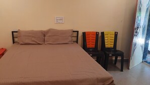 Une pièce comprenant un lit, deux chaises, une petite table rouge et une prise de courant au mur.