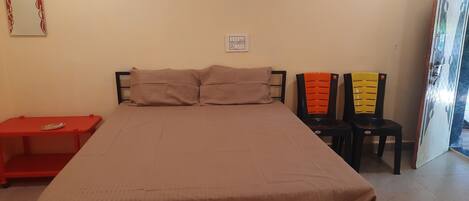 Une pièce comprenant un lit, deux chaises, une petite table rouge et une prise de courant au mur.