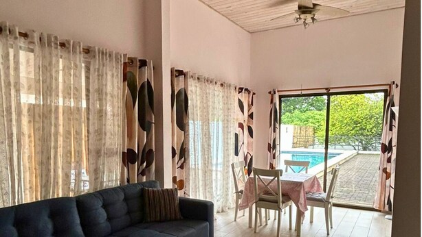 A living room with a dark sofa, a dining table with chairs, and a view of a swimming pool through large windows. - Casa Terracota: Lovely 3-bedroom house in Atenas Perfect for Relaxing Getaways (Atenas)