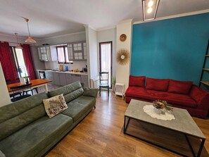 A living room with a green sofa, a red sofa, and a coffee table. There is a kitchen in the background with grey cabinets and a window with curtains. - My Apartments Tirana (Tirana)