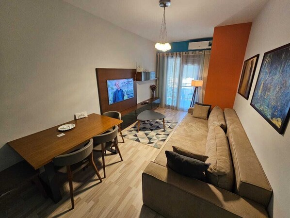A modern living room with a wooden dining table, a sofa, and a TV mounted on the wall. - My Apartments Tirana (Tirana)