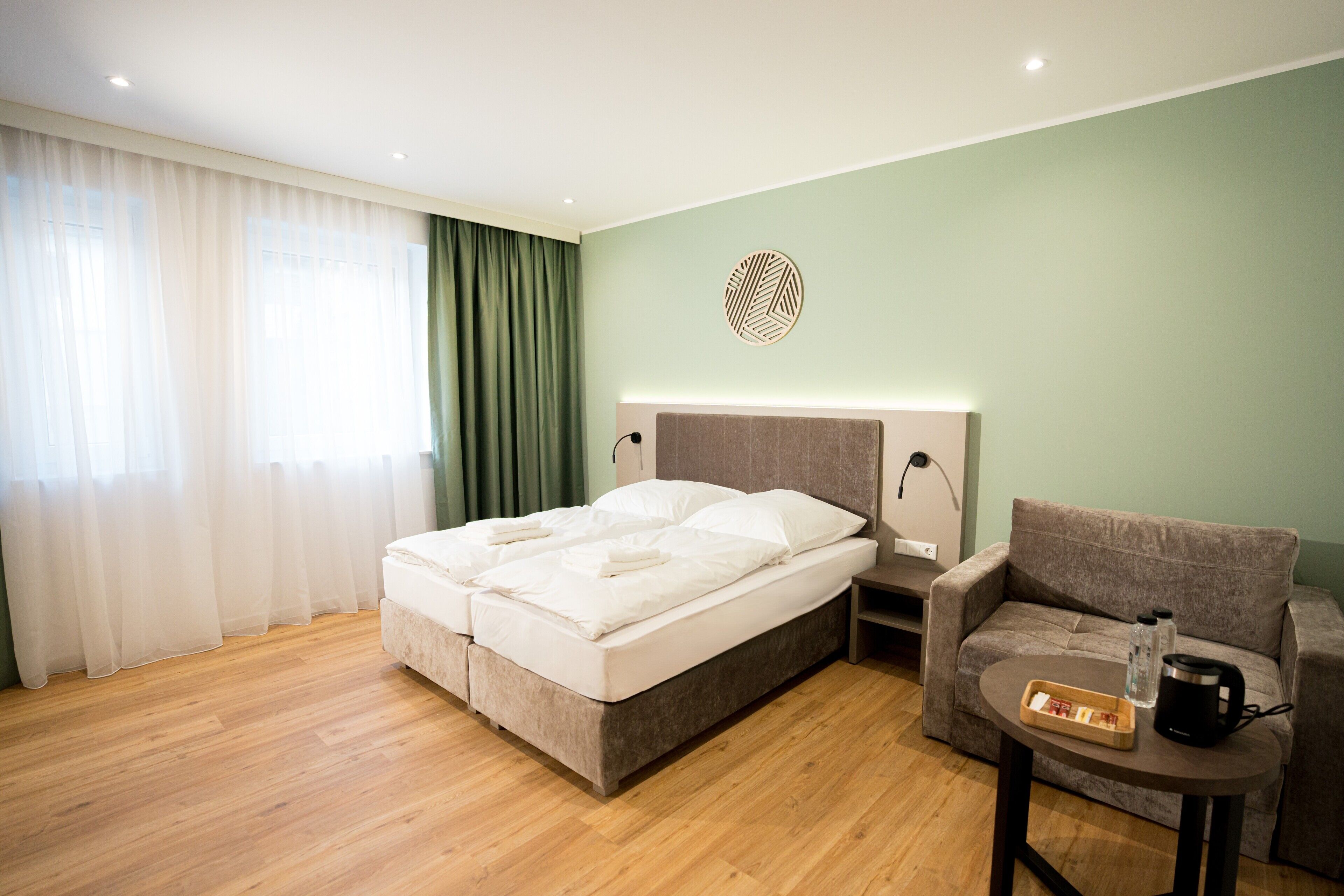 A modern hotel room with a bed, a sofa, a small table, and a wall-mounted light fixture.