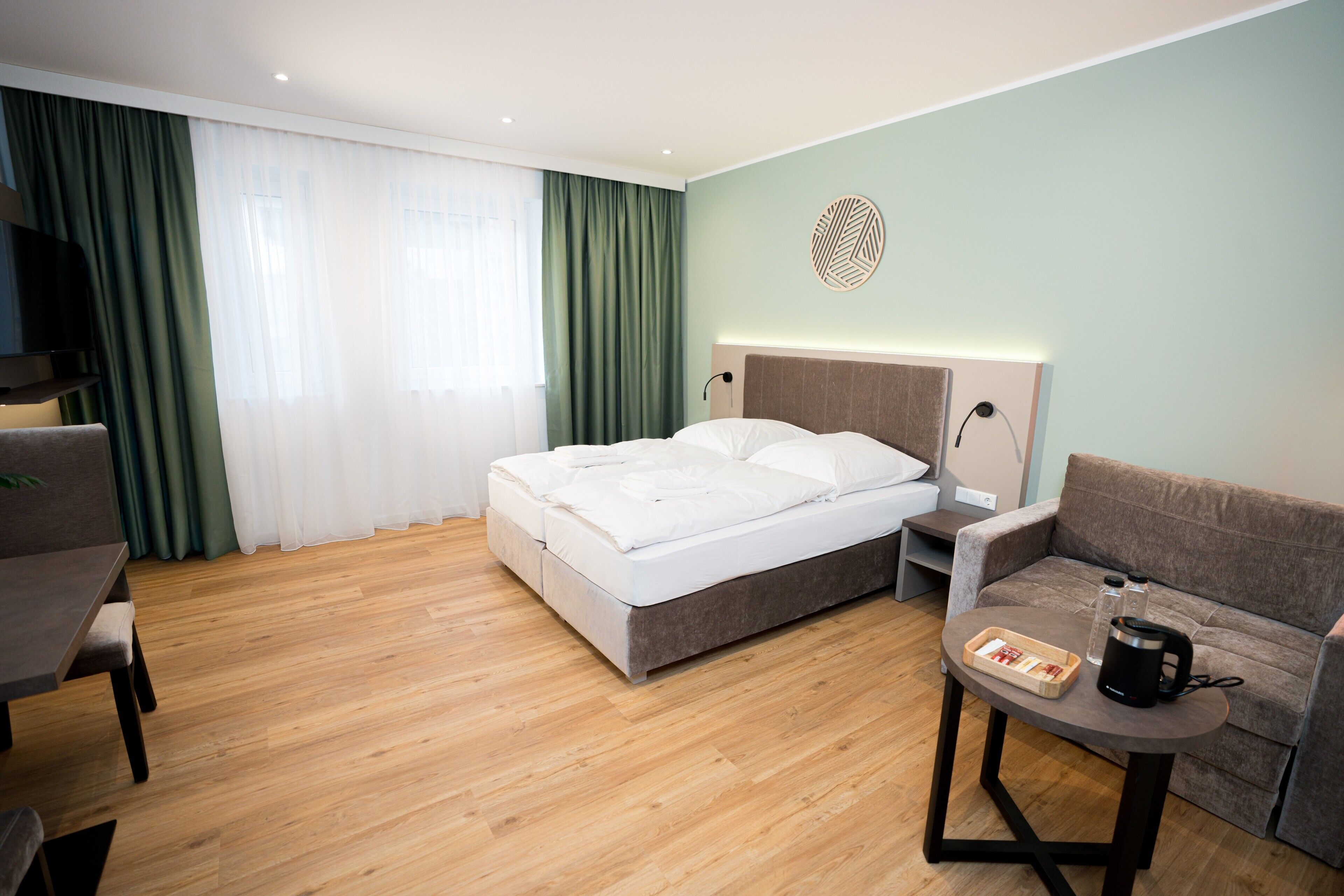 Una habitación de hotel con cama, sofá, una pequeña mesa con tetera y vasos, televisor y ventana con cortinas.