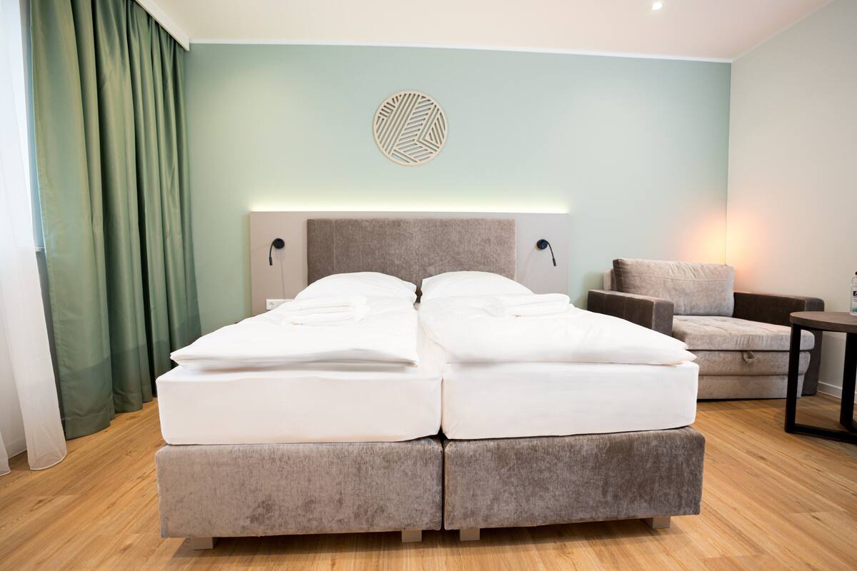 Un lit double avec une tête de lit, une chaise, une petite table et un luminaire fixé au mur.