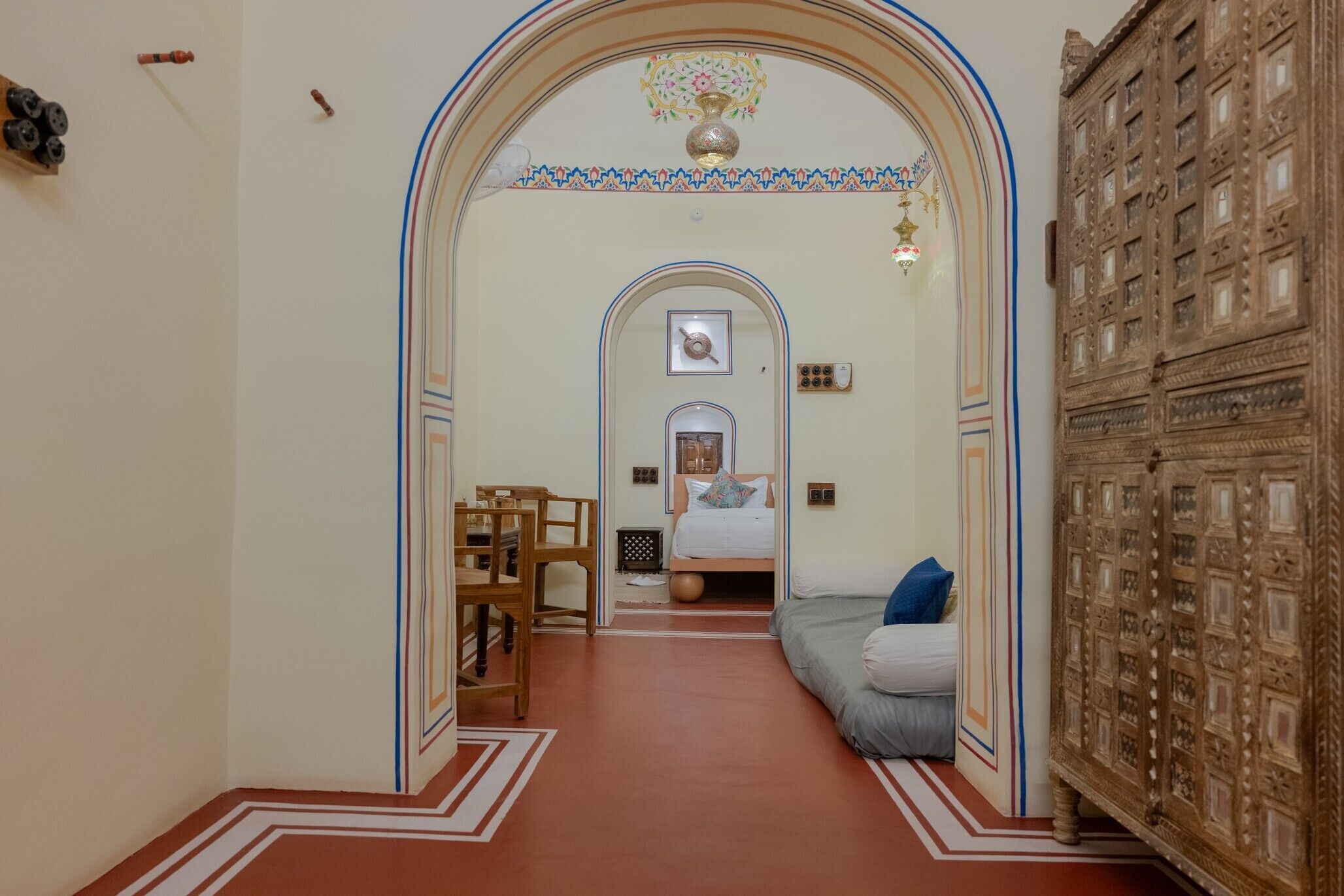 A hallway leading to a bedroom with an ornate wooden cabinet, a bed with a blue pillow, and a small table with chairs.