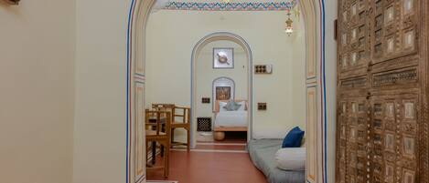 A hallway leading to a bedroom with an ornate wooden cabinet, a bed with a blue pillow, and a small table with chairs.