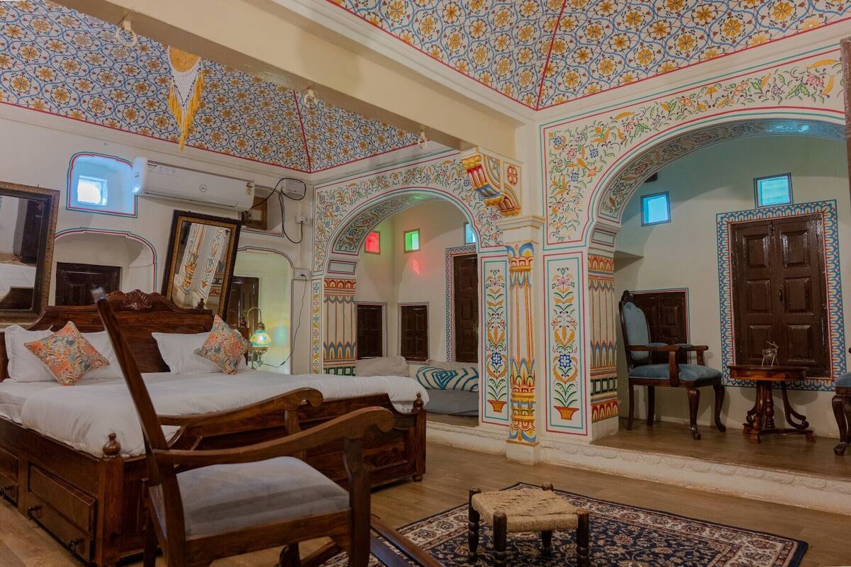 Una habitación tradicional con una cama de madera, una mesita pequeña y una silla. La habitación presenta motivos florales ornamentales en las paredes y el techo.