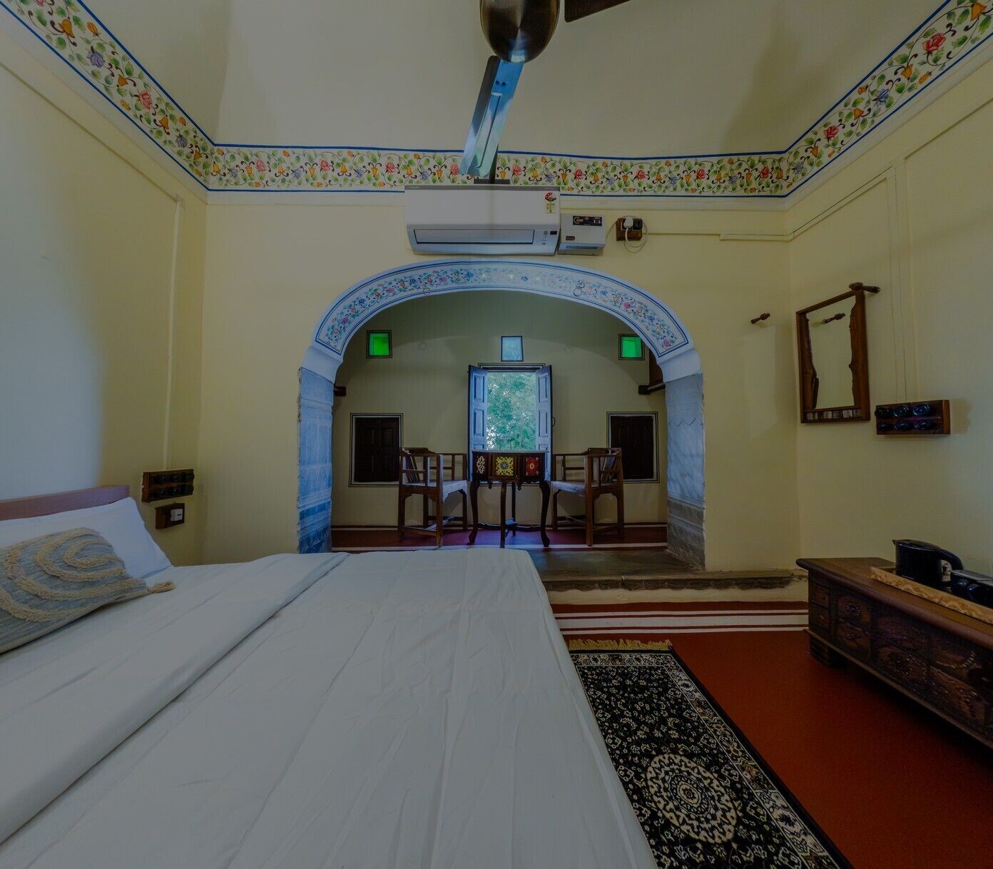 A room with a bed, a desk with chairs, an air conditioning unit, and a mirror on the wall.
