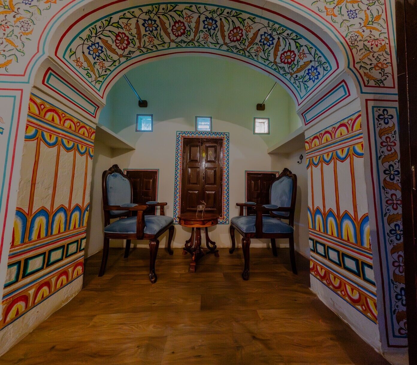 Una habitación con dos sillas de madera, una mesita pequeña y una puerta grande de madera. Las paredes están decoradas con motivos coloridos.