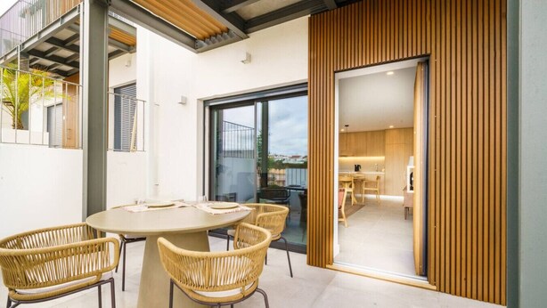 A modern outdoor dining area with a round table and wicker chairs, a glass door leading to a kitchen, and a view of the surrounding area. - Carvoeira Nature & Serenity Escape by Lisbeyond (Barril)