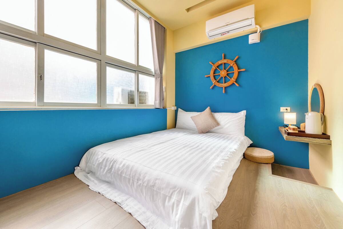 Un dormitorio con un ventanal grande, una cama con ropa de cama blanca, una pared azul de acento, un adorno de timón de barco y una mesita de noche con una lámpara.