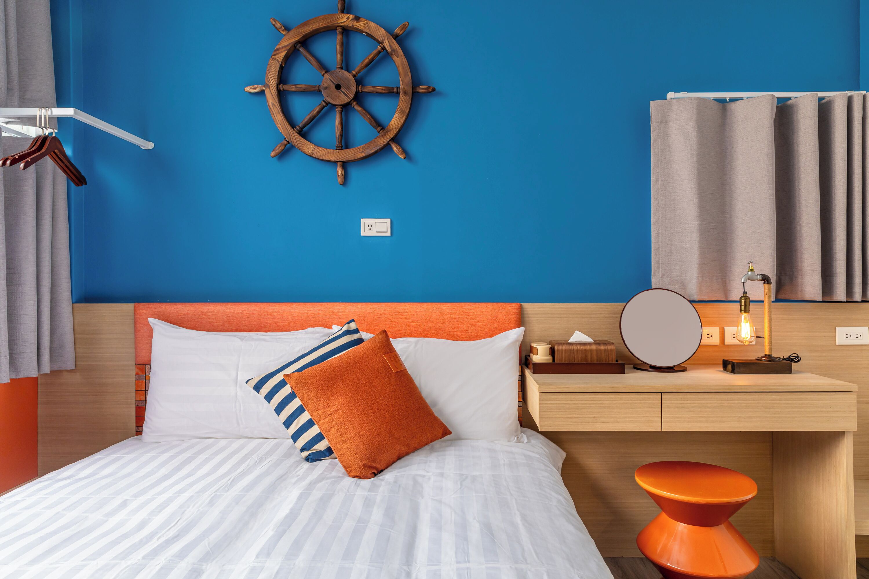 Una cama bien hecha con almohadas a rayas y lisas, una rueda de barco de madera en la pared, una mesita de noche con lámpara y un taburete naranja moderno.