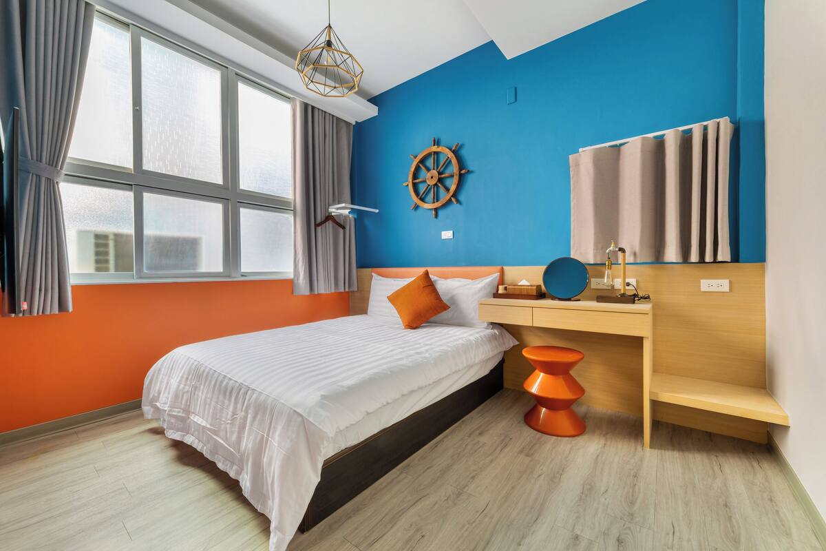 Un dormitorio moderno con una cama grande, un escritorio de madera, un adorno con timón de barco y una ventana con cortinas.