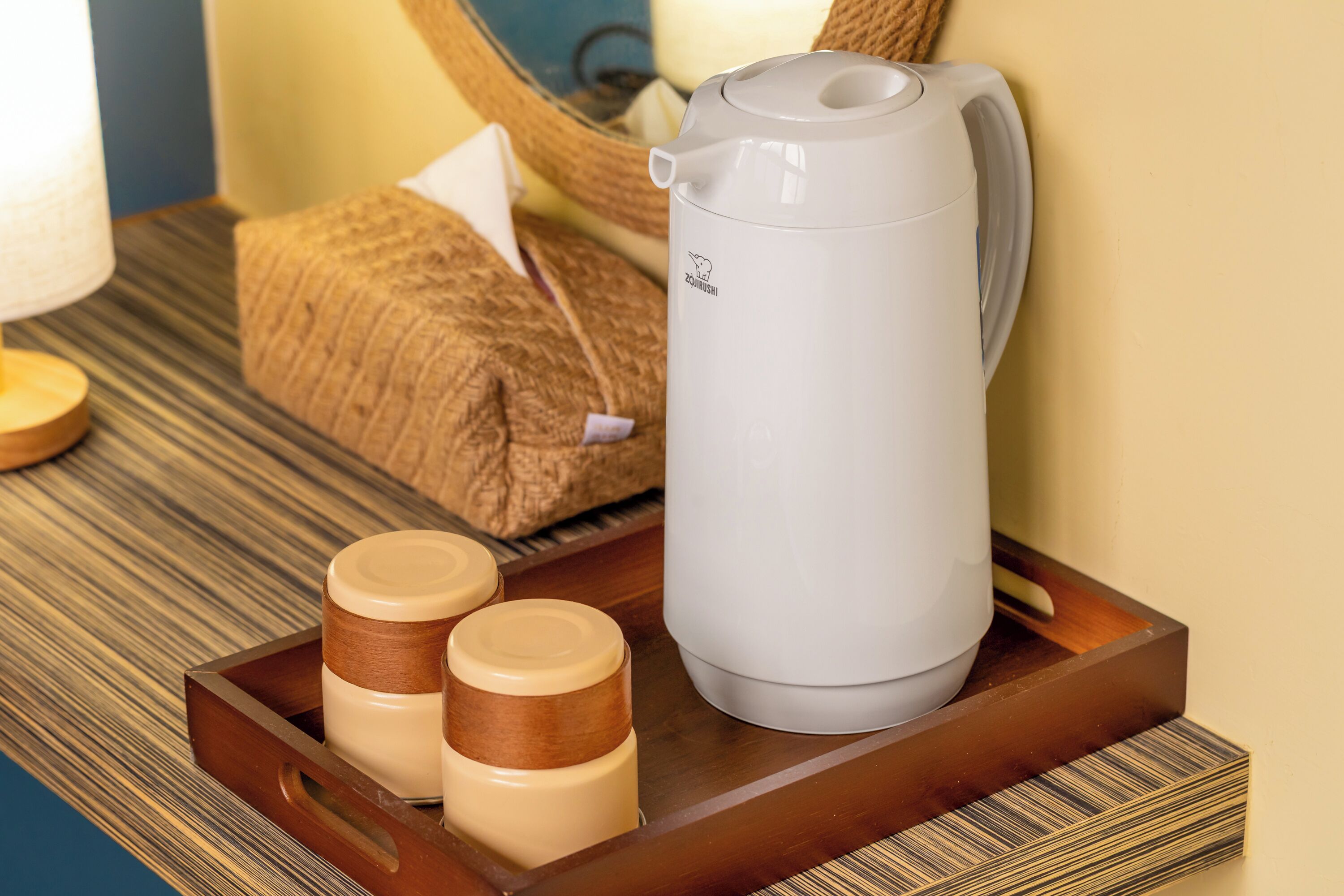 A white electric kettle on a tray with two containers, a woven bag, and a wooden table with a striped pattern.