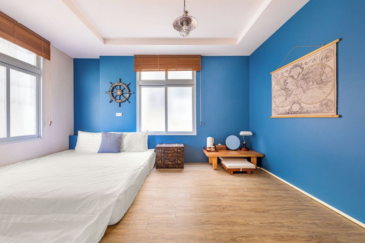Un dormitorio con una cama grande, un cofre de madera, un escritorio con lámpara, un mapa en la pared y una ventana con persianas.