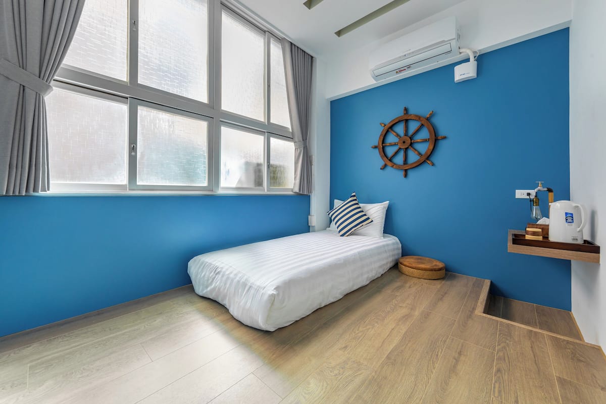 Una habitación con cama, ventana, timón de barco en la pared y una estantería con hervidor.