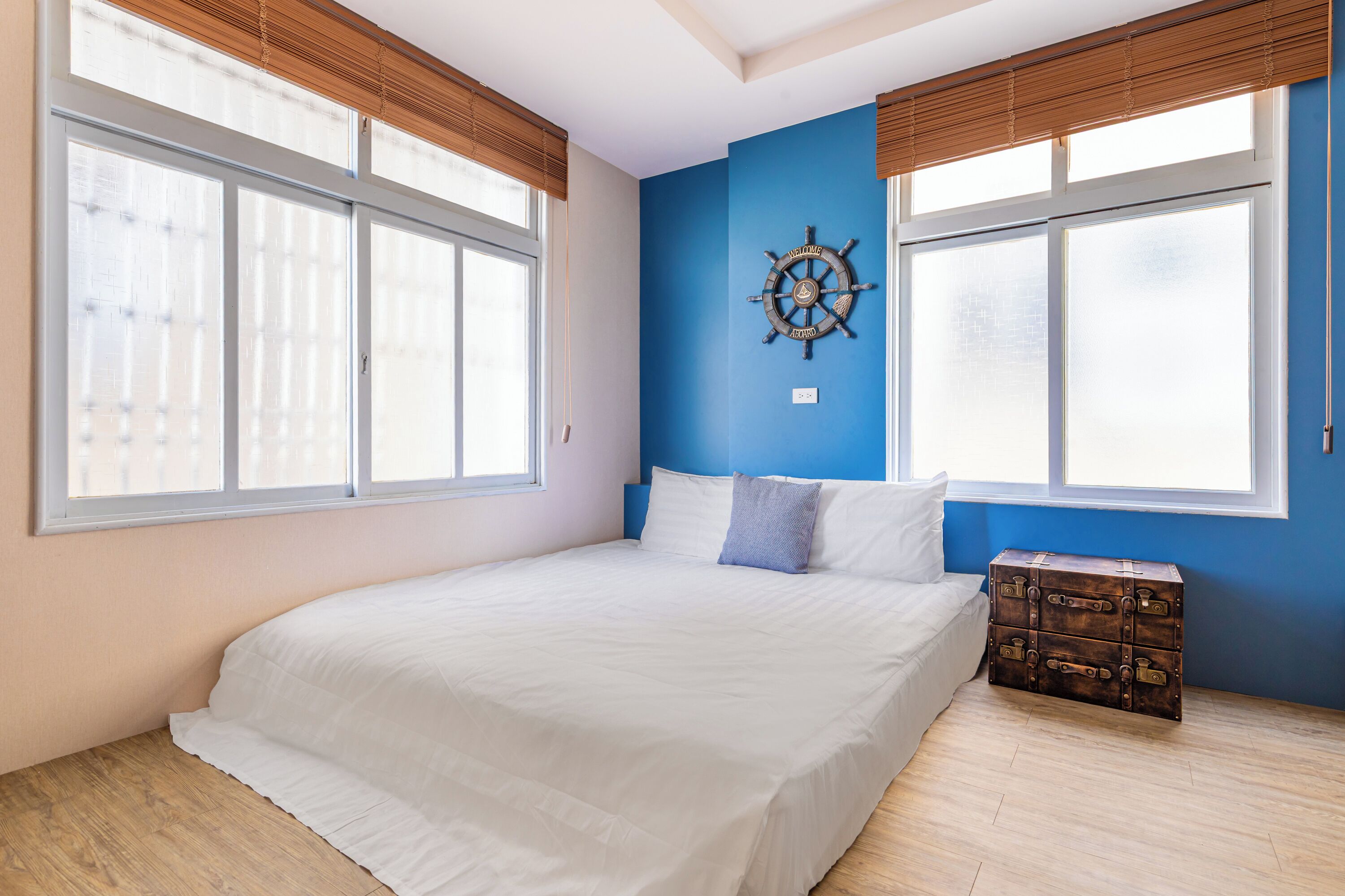 A bedroom with a large bed, a wall-mounted ship's wheel decoration, and a wooden chest of drawers.