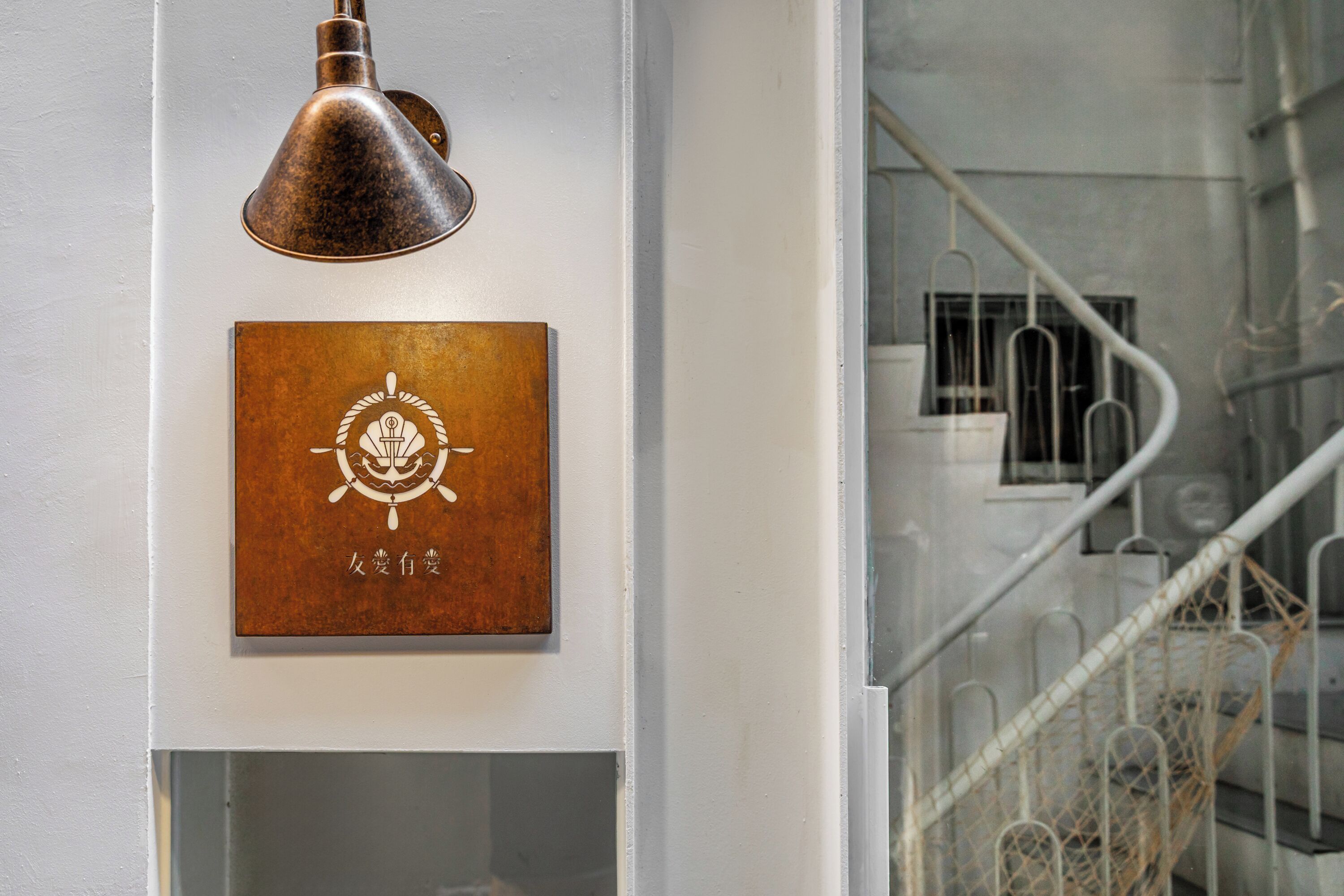 En vægplade med et dekorativt emblem og tekst, en ståltrappe med gelænder, og en vægmonteret lampe.