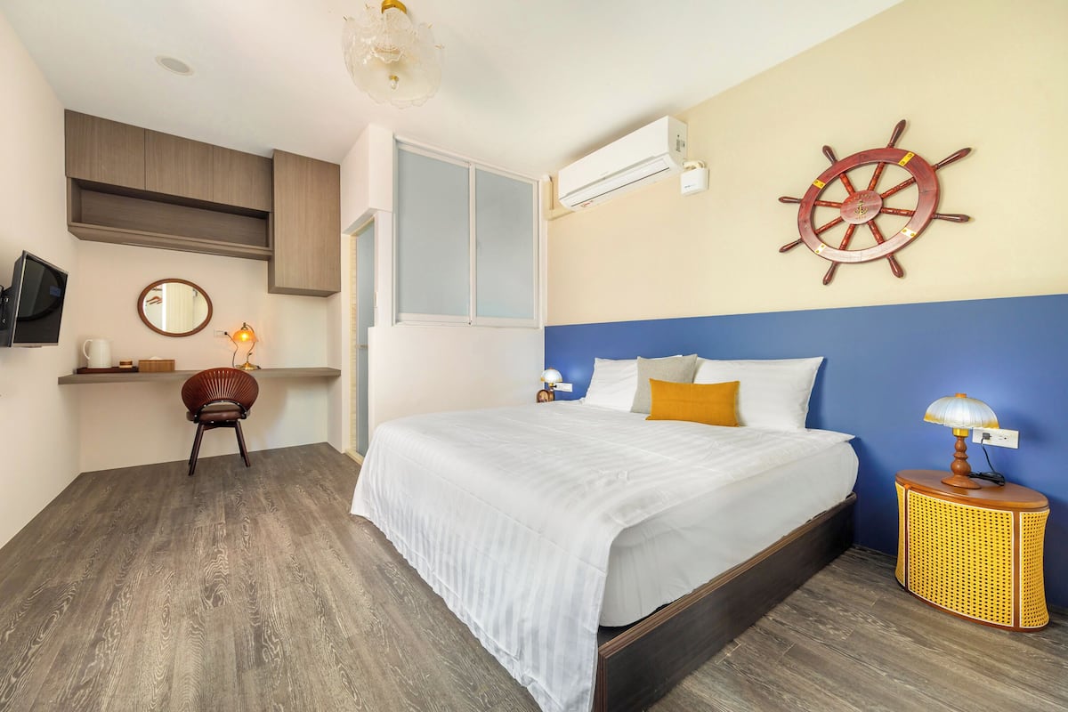 Un dormitorio con cama, una silla de madera, una mesita de noche con lámpara, un timón de barco en la pared y un equipo de aire acondicionado.