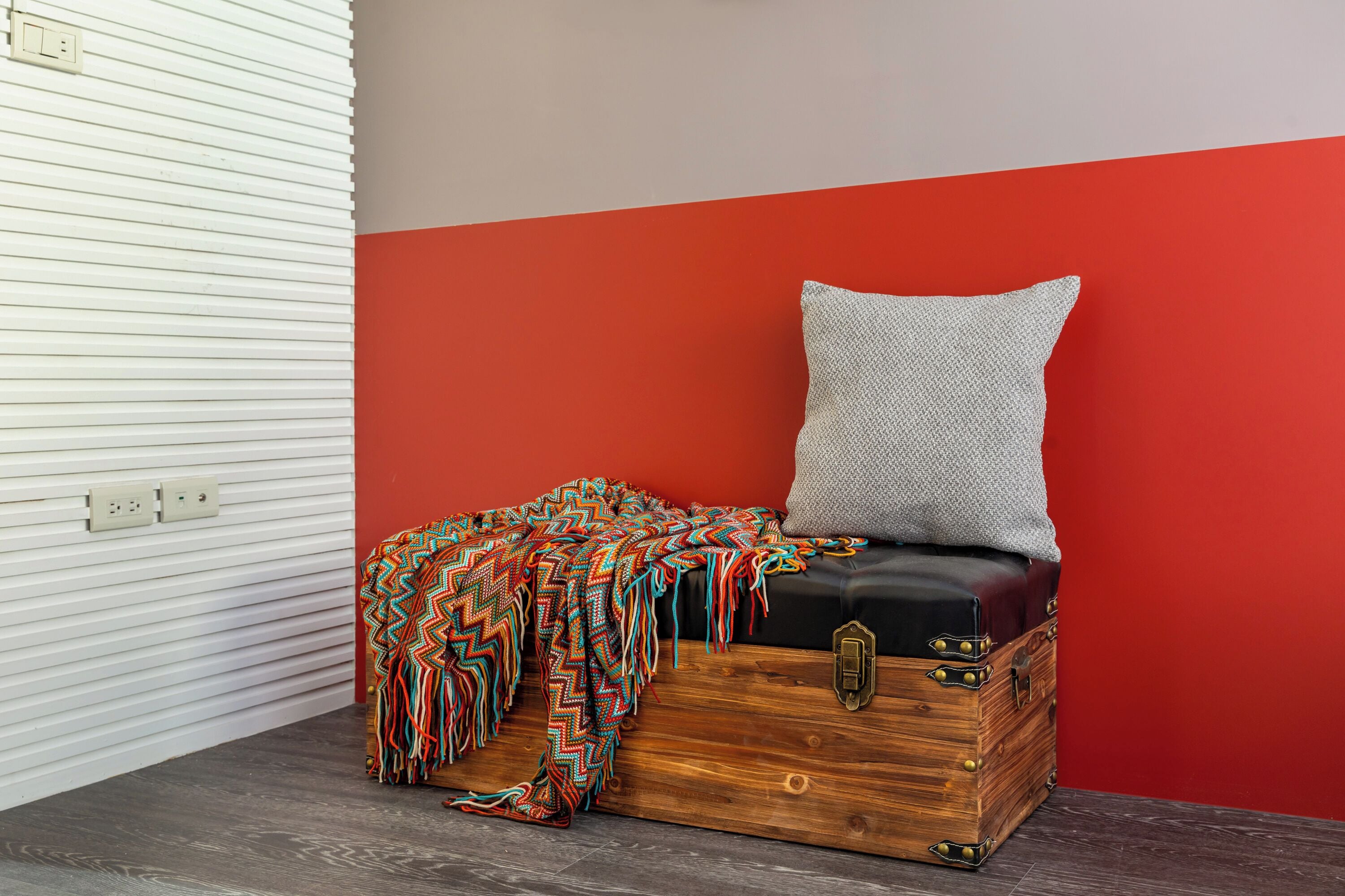 A room with a red wall, a wooden chest with a cushion, and a colorful blanket.