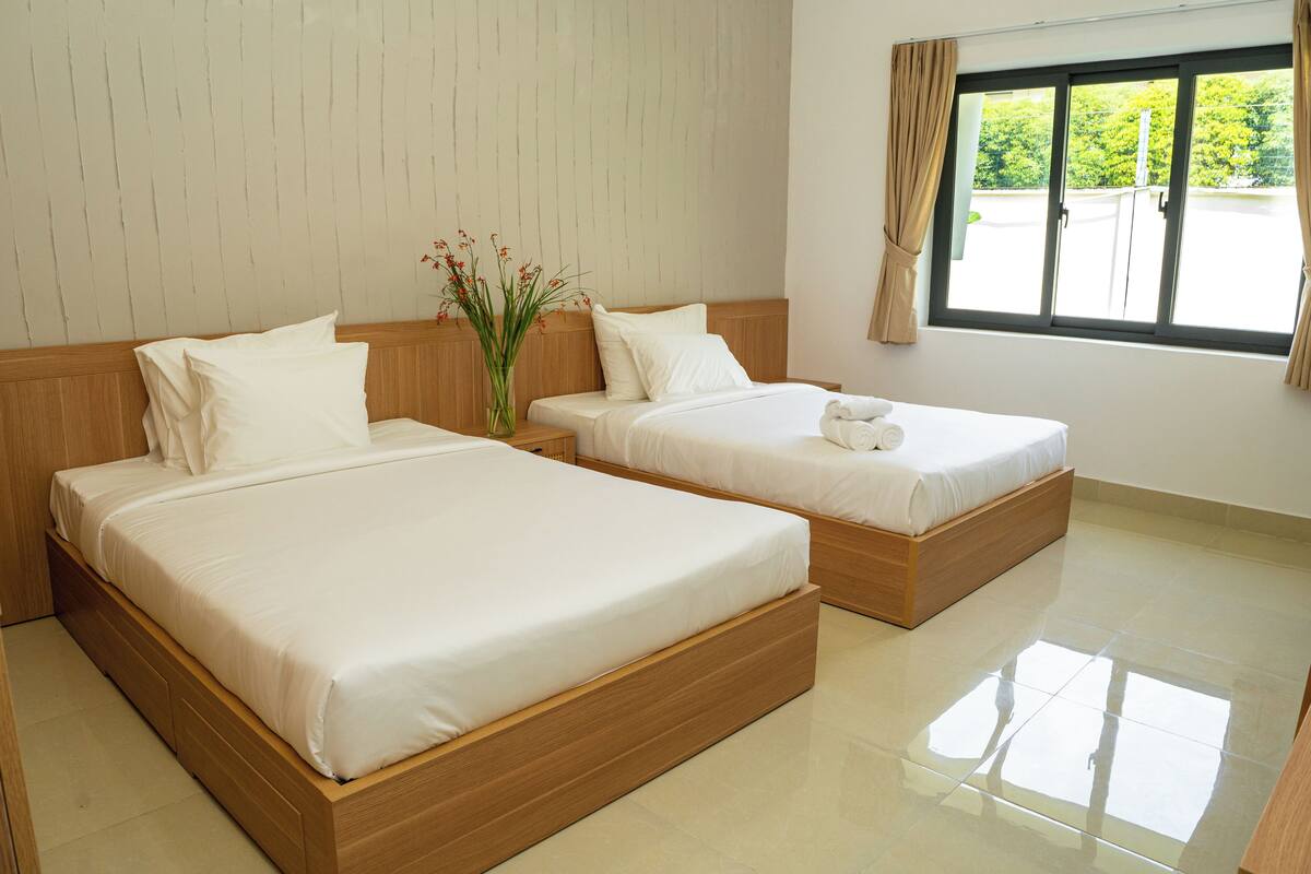 Quarto de hotel com duas camas de madeira, uma mesinha de cabeceira, um vaso com flores e uma janela com cortinas.