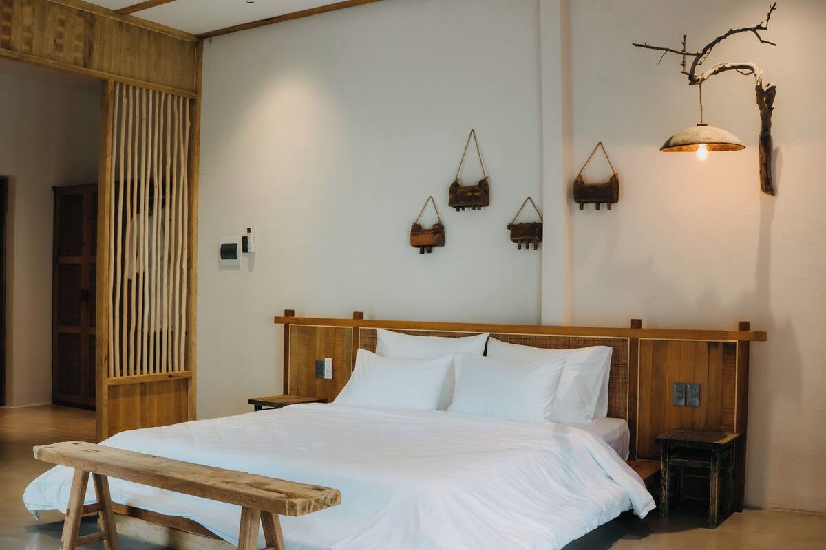 Quarto com cabeceira de madeira, cama com roupa de cama branca, mesa de cabeceira, um banquinho e objetos decorativos fixados na parede.