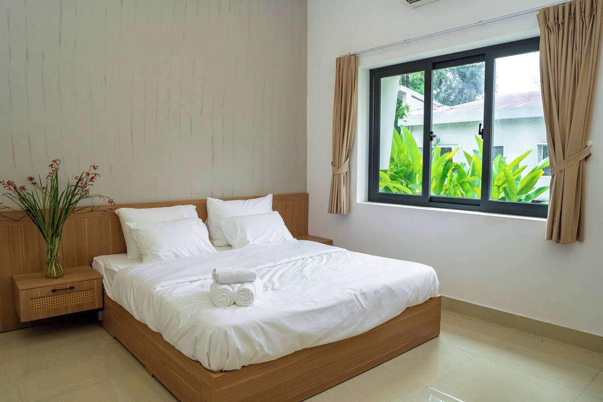 Cama bem arrumada com lençóis brancos, cabeceira de madeira, mesa de cabeceira com um vaso de flores e janela com cortinas.