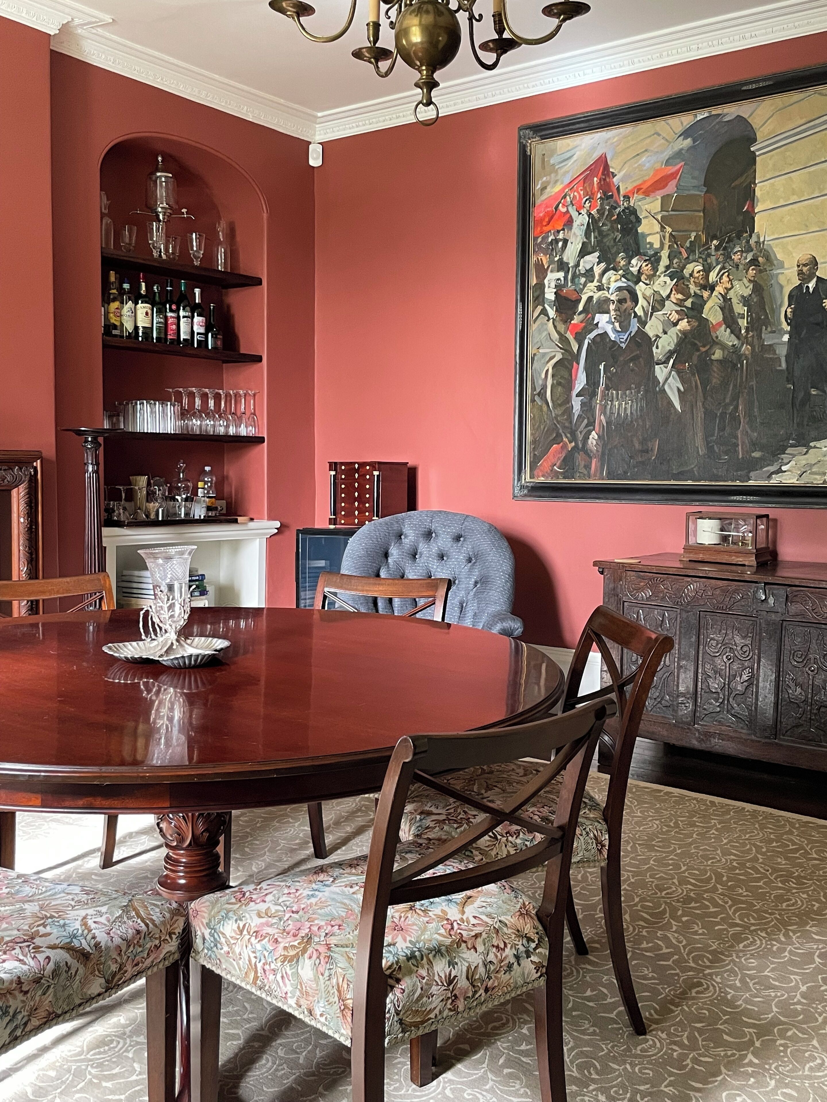 Une salle à manger avec une grande table en bois, des chaises rembourrées, un lustre, un meuble à vin et un grand tableau accroché au mur.
