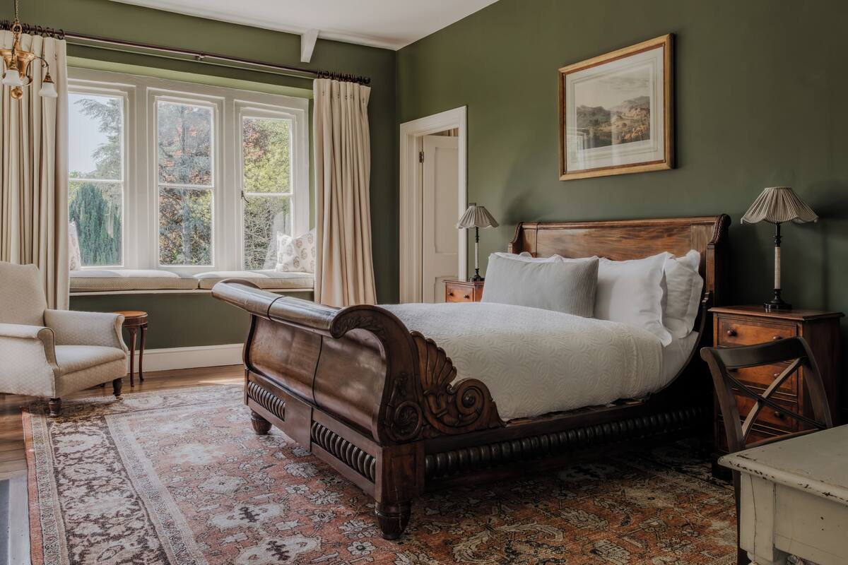 A bedroom with a large bed, a window seat, a chair, a bedside table, and a lamp.