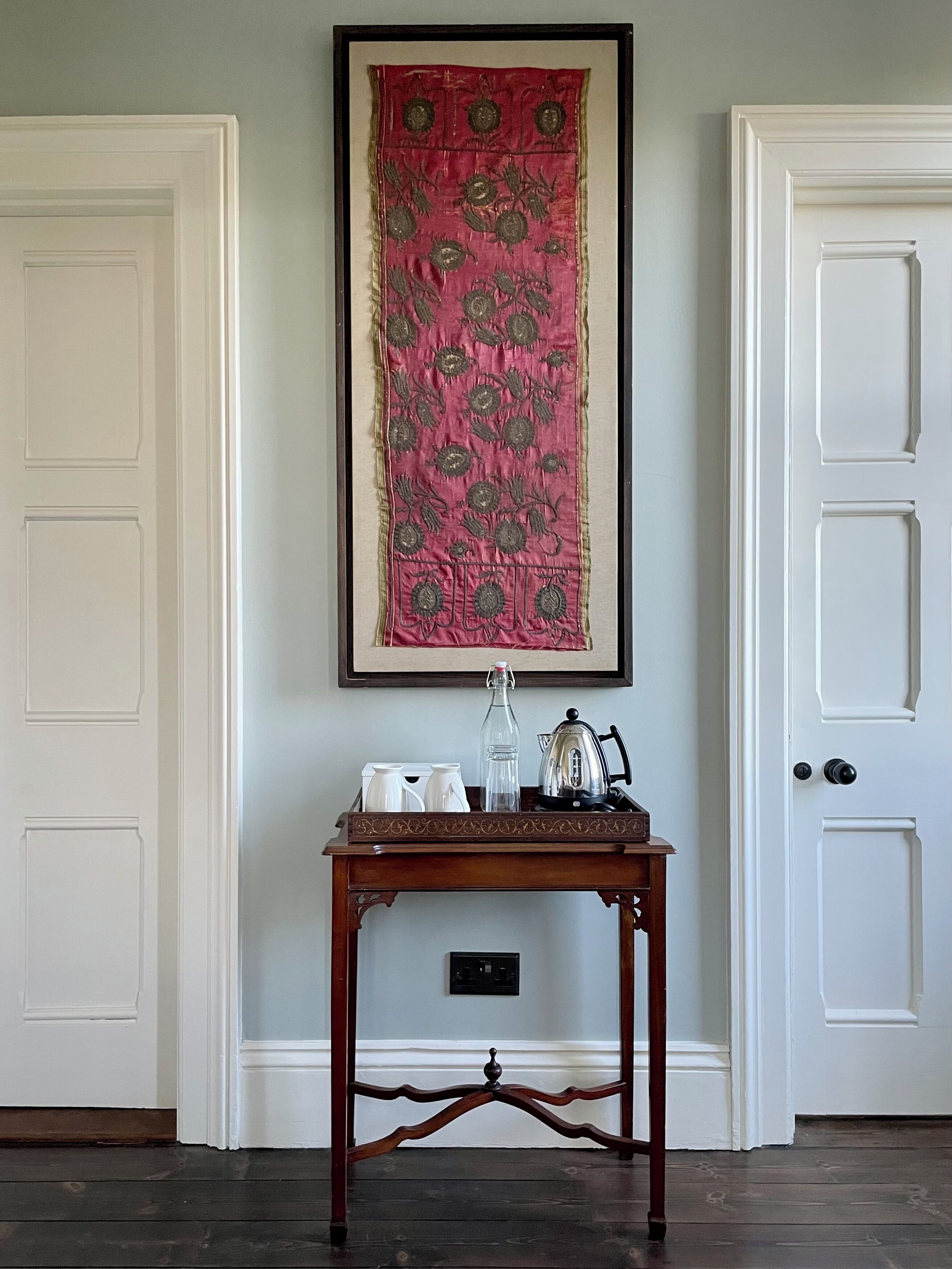 A room with a white door, a framed tapestry, a small wooden table with a tray holding a kettle, cups, and a bottle.