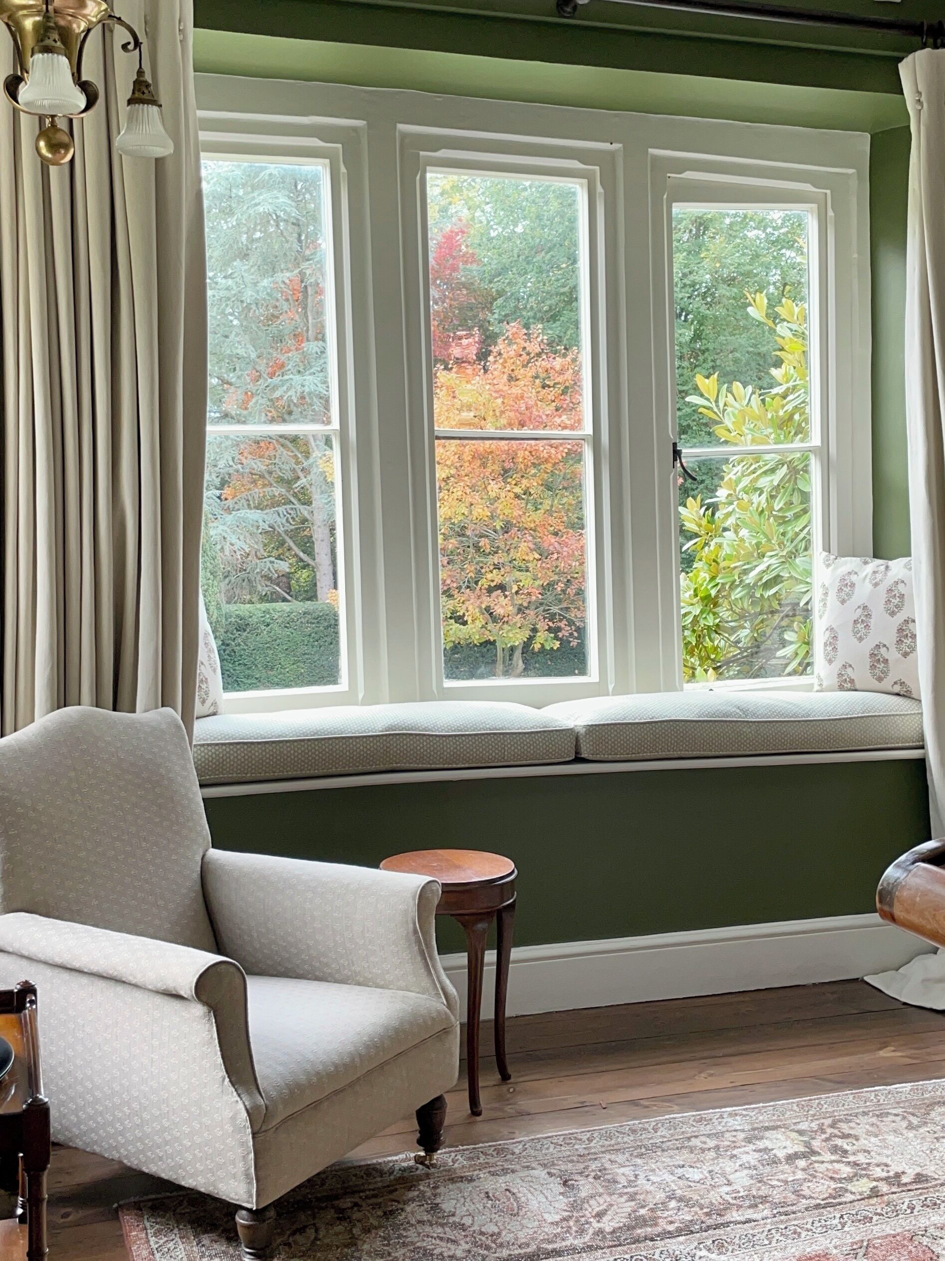 A room with a large window, a cushioned bench, a chair, and a small table.