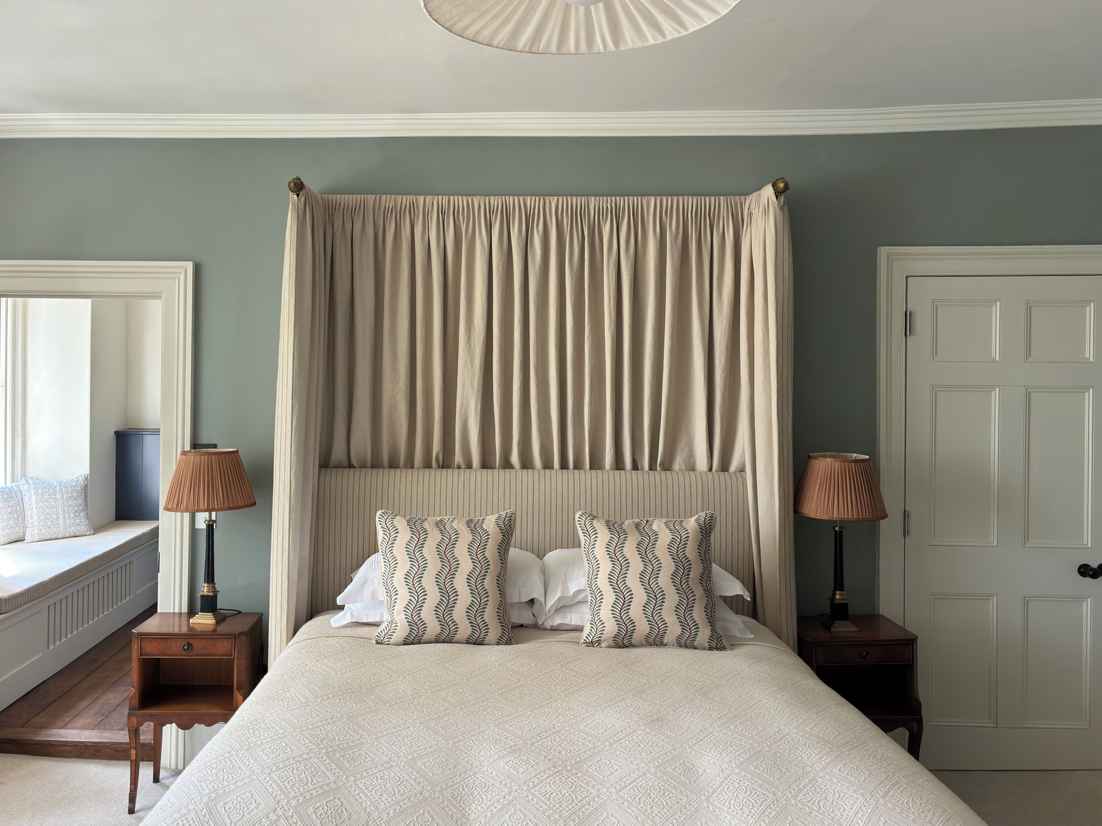 Un dormitorio con una cama grande, dosel, dos mesitas de noche con lámparas, ventana con vistas y una puerta.