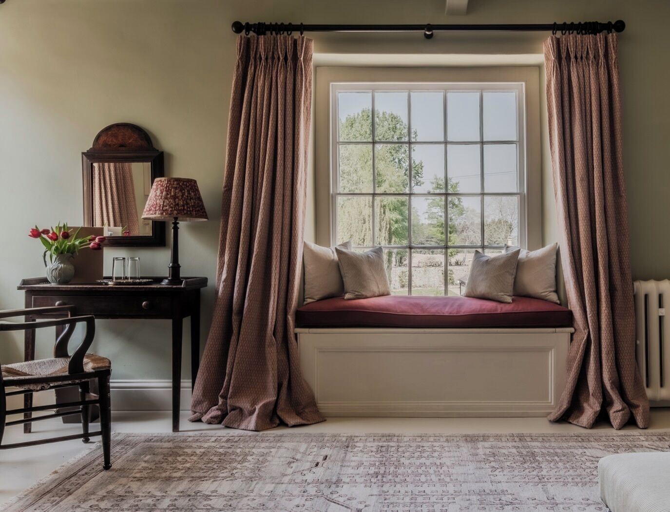 A room with a window seat, a desk, a chair, a lamp, and a mirror.
