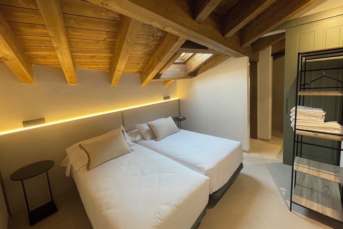 A bedroom with two beds, a bedside table, a hanging shelf, and a sloped ceiling with wooden beams.