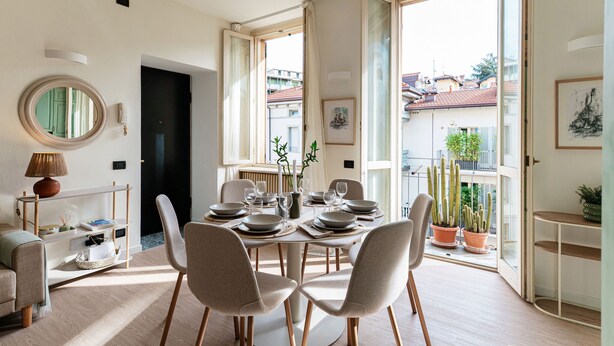 Un espace repas moderne avec une table ronde pour quatre personnes, entourée de chaises. Une fenêtre offre une vue sur les bâtiments, et un balcon est agrémenté de plantes en pot.