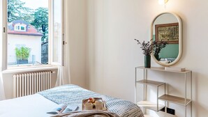 Une chambre à coucher avec un lit, une fenêtre, un radiateur, un miroir et une étagère sur laquelle se trouve un vase.