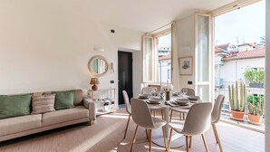 Un salon moderne comprenant un canapé, une table à manger, des chaises et une grande fenêtre donnant sur des immeubles et agrémenté d’un cactus.