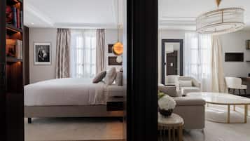 A modern hotel room with a large bed, a sitting area with a sofa and armchairs, a small round table, and a bookshelf.