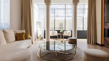 A modern living room with a glass coffee table, a sofa, and two chairs. There are large windows with a view of the city.