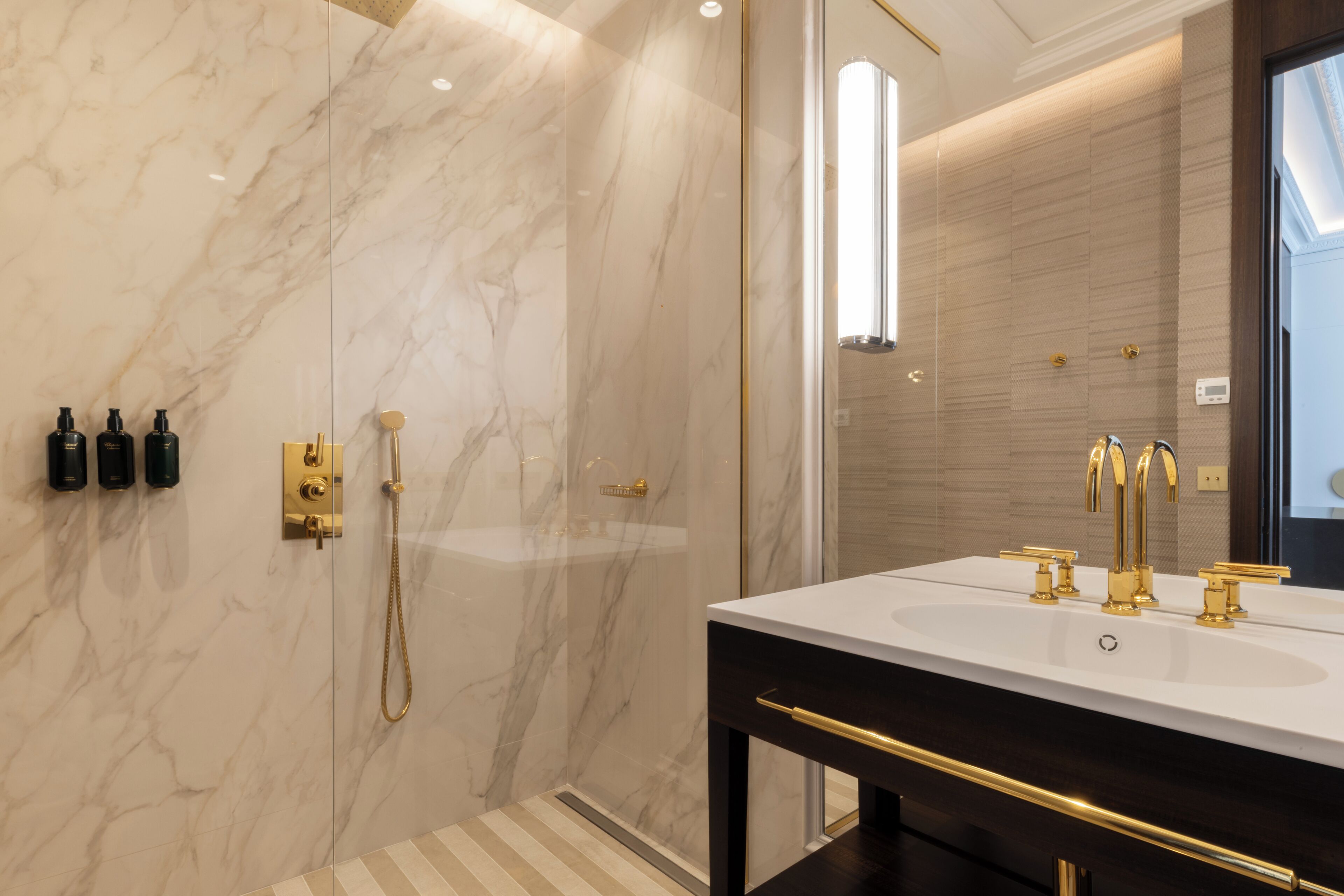 Un baño moderno con pared de mármol, un espejo grande, un lavamanos blanco con detalles dorados y zona de ducha.