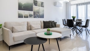 A modern living room with a sofa, a coffee table, and a dining area with chairs and a table. There are wall-mounted air conditioning units and framed pictures on the wall. - Averna - Breezy 2-BR Apt. in Larnaca (Larnaca)