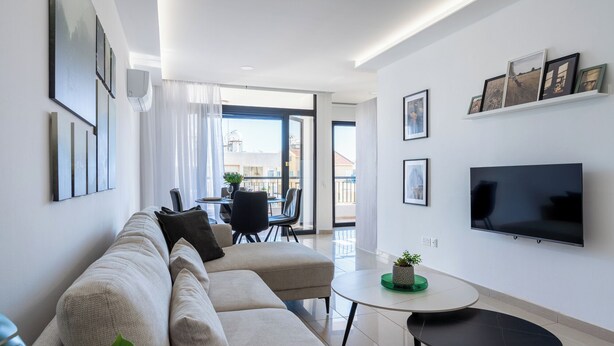 A modern living room with a sofa, a TV, and a dining area with a table and chairs. - Averna - Breezy 2-BR Apt. in Larnaca (Larnaca)