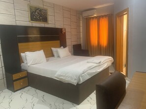 A bedroom with a large bed, a nightstand, a chair, a wall-mounted air conditioner, and a door. - Comfort Kul Suites (Agbor)