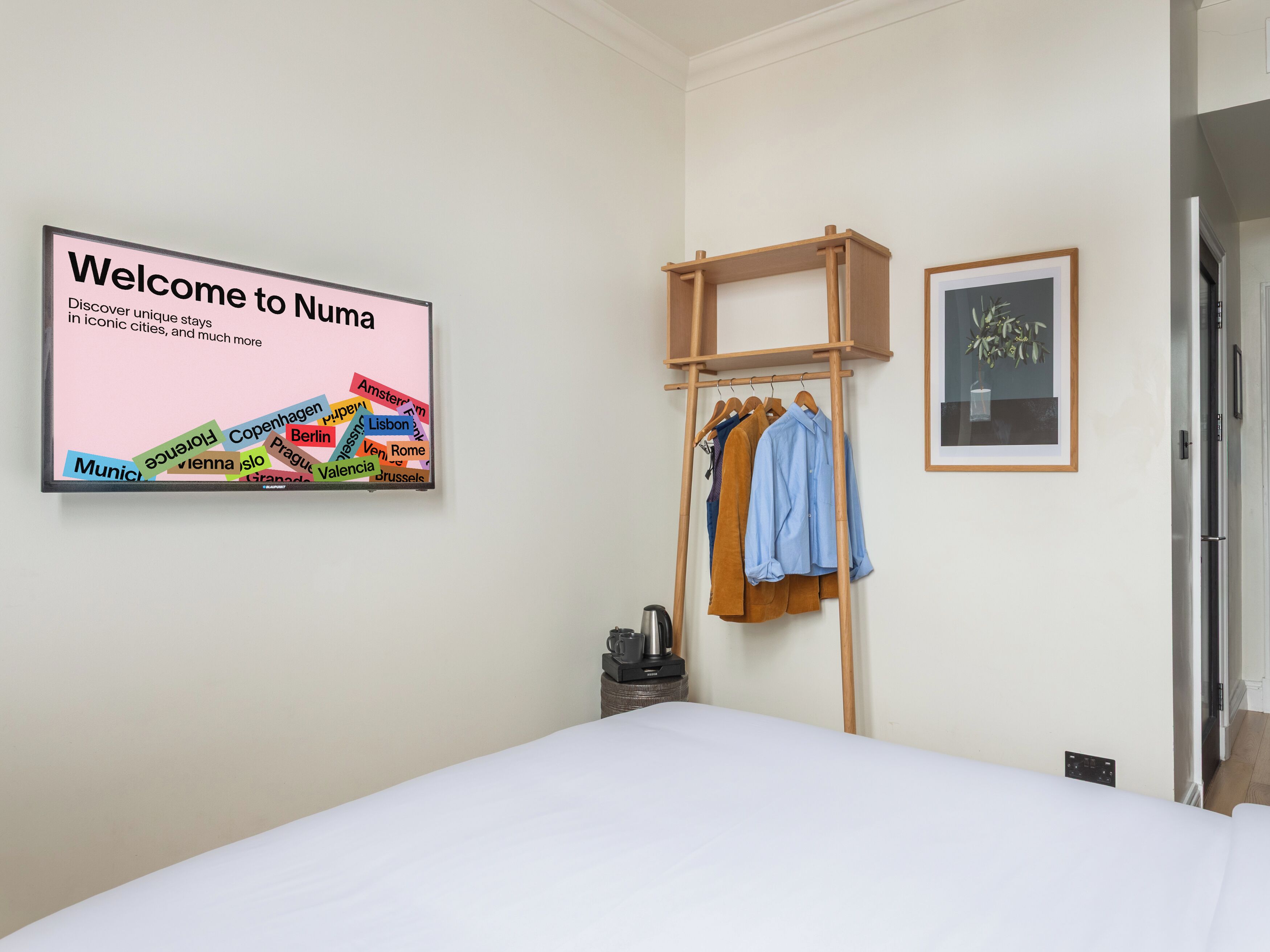 A hotel room with a bed, a wall-mounted sign with city names, a wooden coat rack with clothes, and a framed picture.