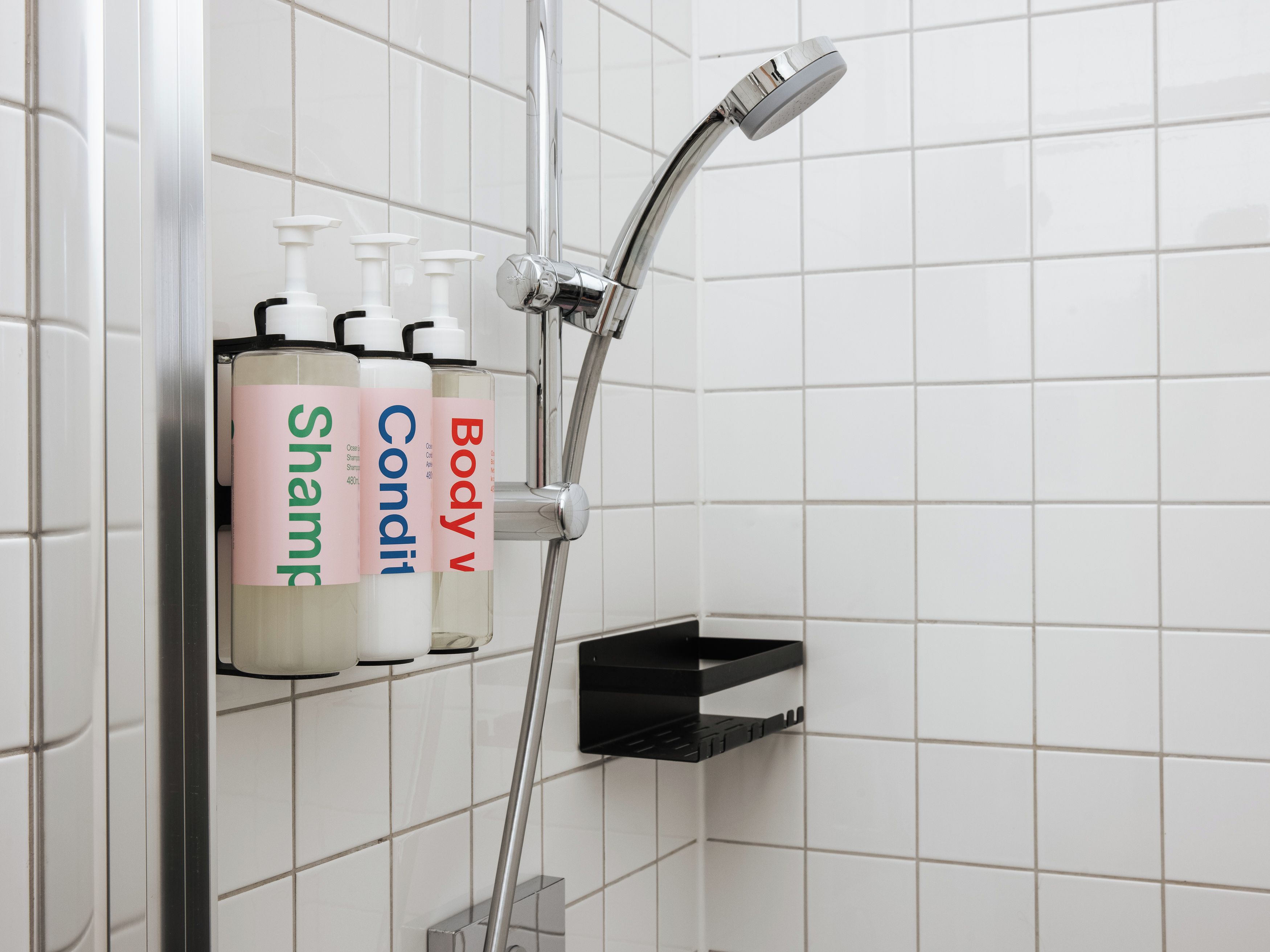 A bathroom with a shower, a black shelf, and three bottles of shampoo labeled 'Shampoo,' 'Condition,' and 'Body V.'