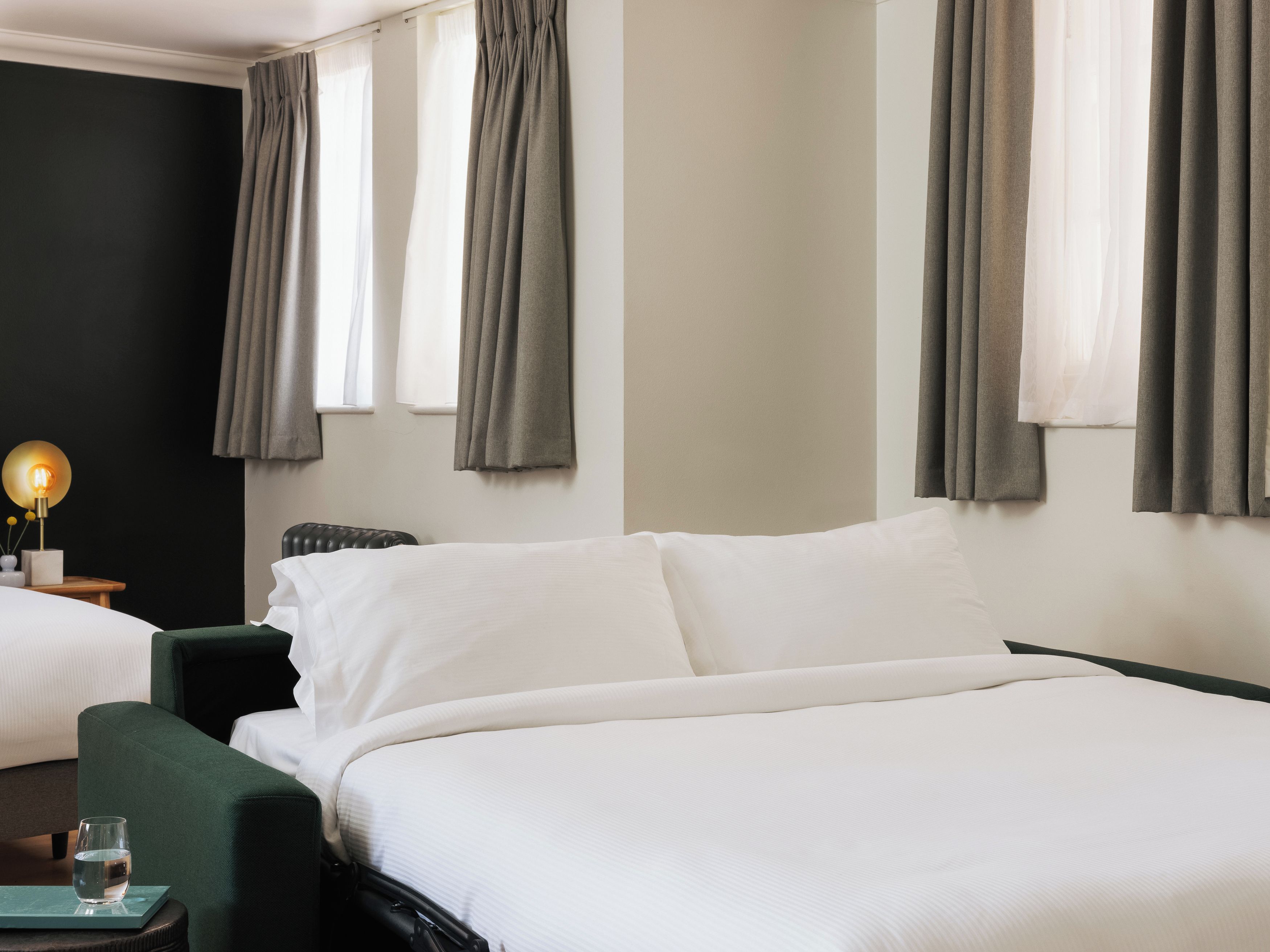 Hotellihuone, jossa on sänky, yöpöytä, lamppu ja ikkuna, jossa on verhot.