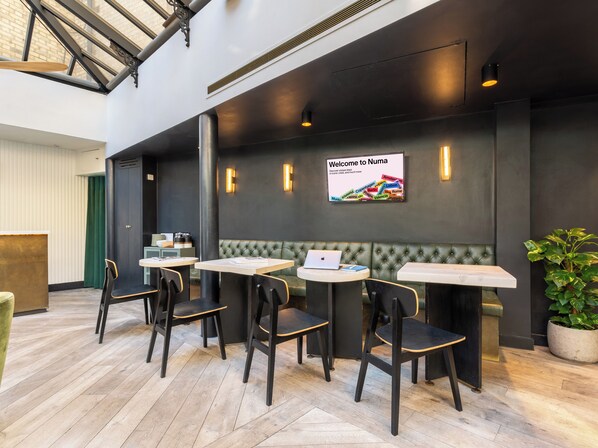 A modern indoor seating area with tables and chairs, a green cushioned bench, and a sign welcoming visitors to Numa. - Numa London Chelsea Green (London)