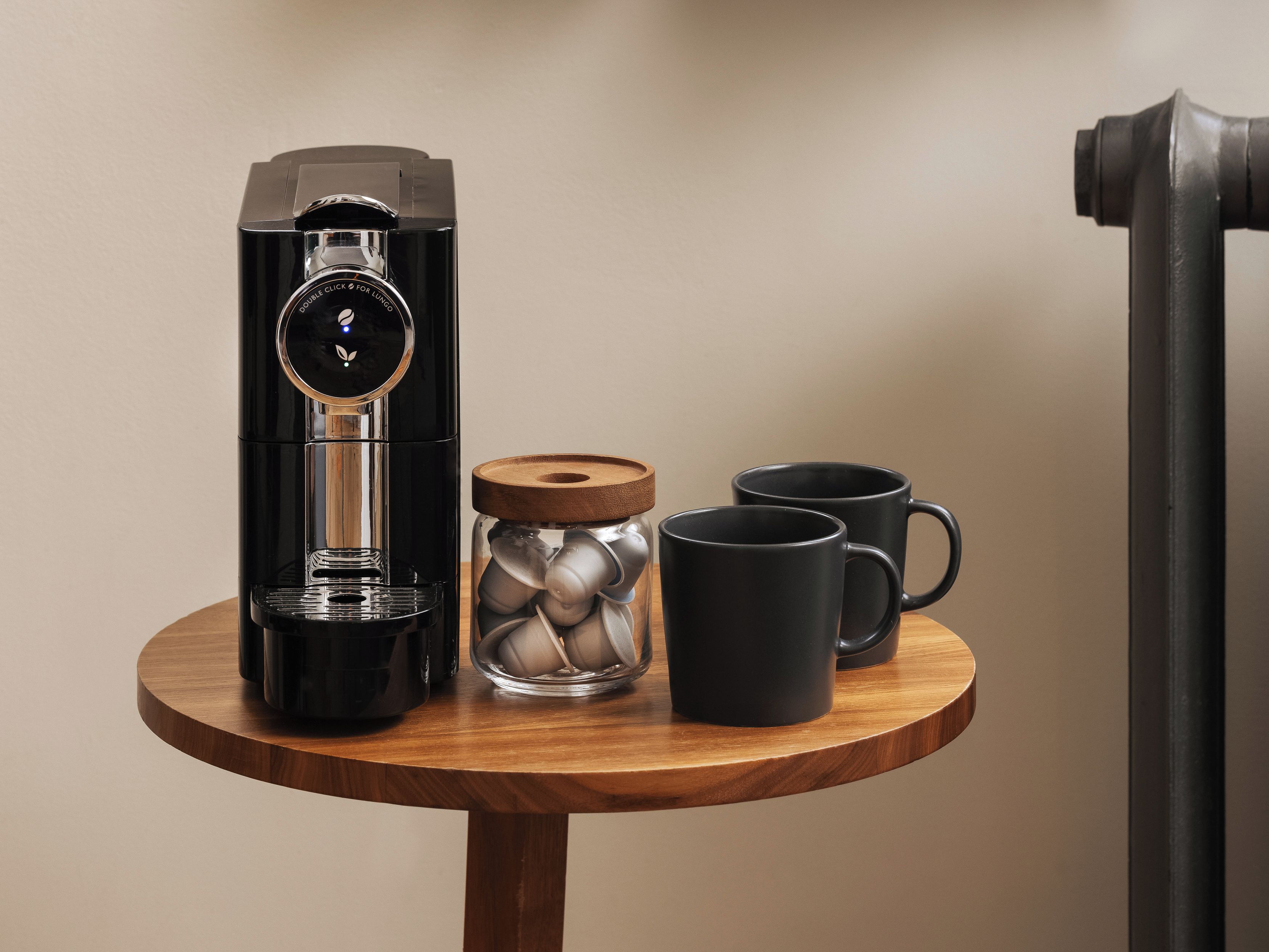 A coffee machine, a glass jar with metal lids, and two black mugs on a wooden table.