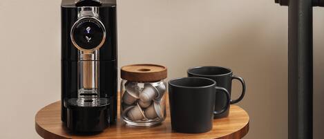 A coffee machine, a glass jar with metal lids, and two black mugs on a wooden table.