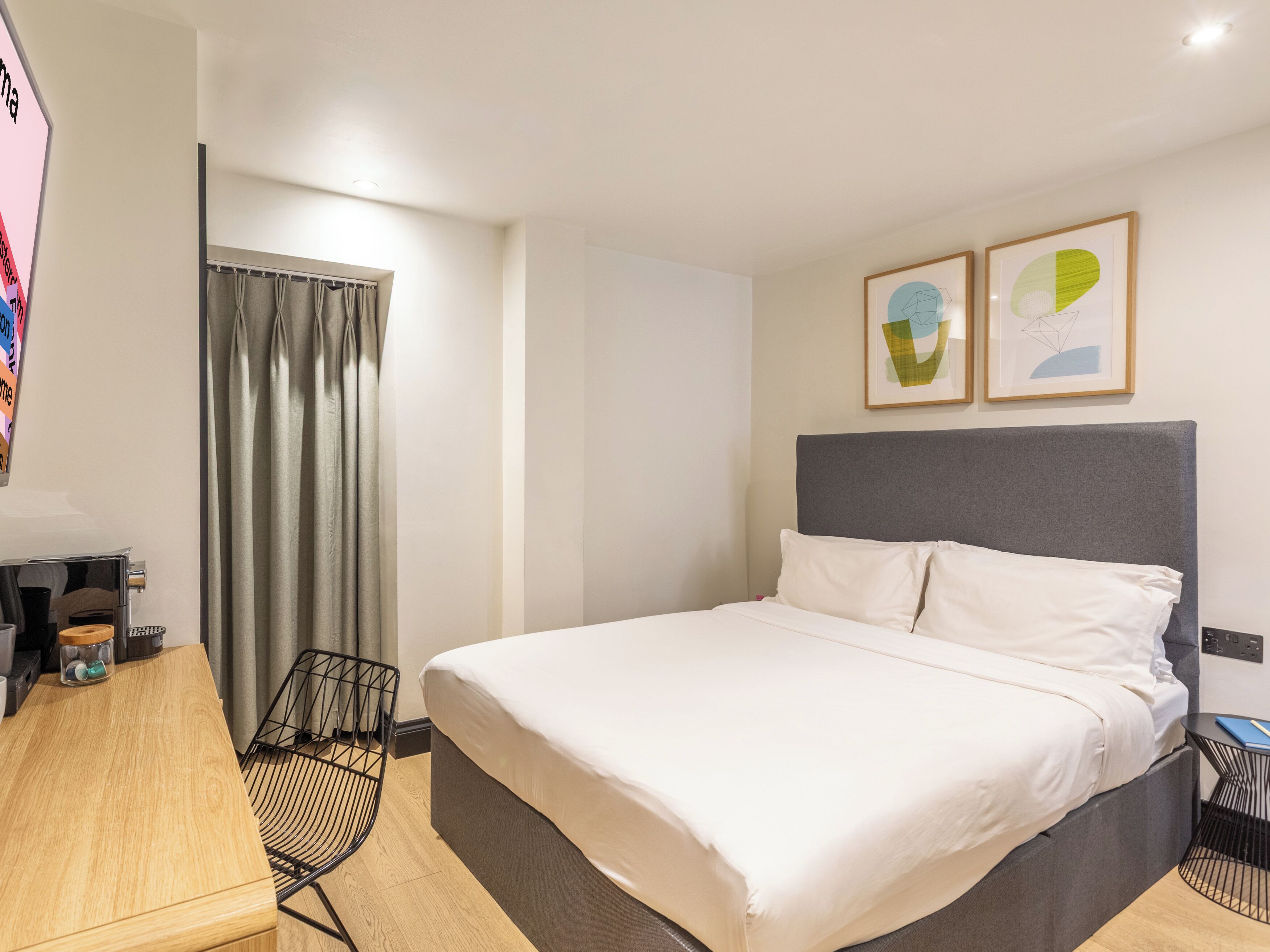 A modern hotel room with a large bed, a desk with a coffee machine, and two framed abstract paintings on the wall.